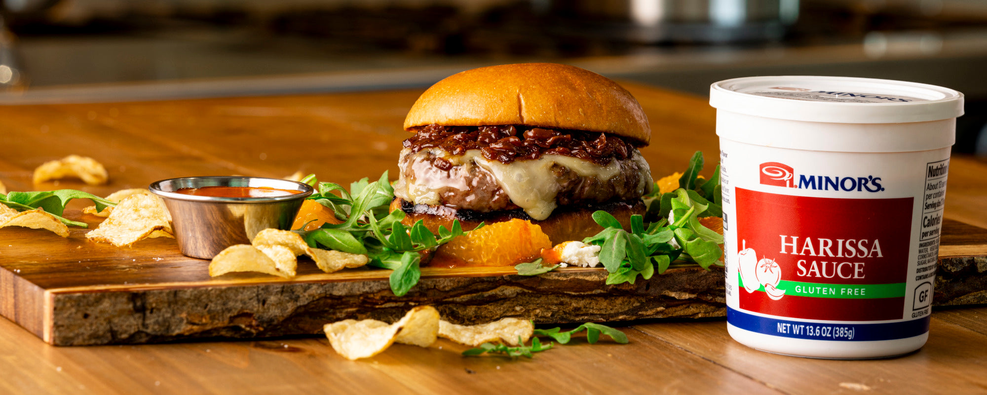 Grilled burger with harissa onion jam on a wooden cutting board with chips and a MINOR'S Harissa sauce container