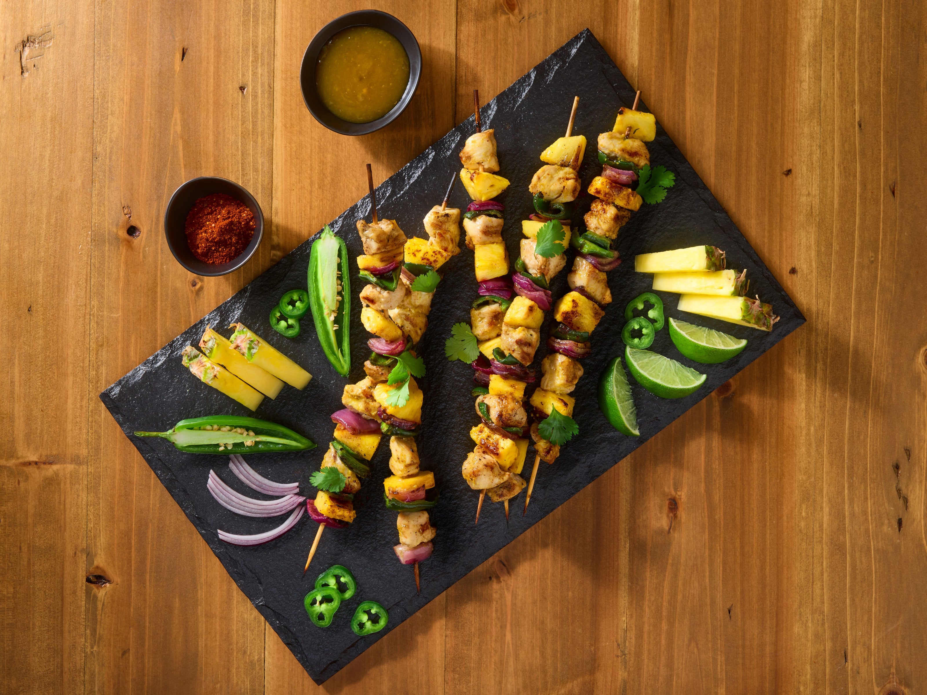Skewers of grilled vegetables and chicken on a dark slate platter with dipping sauces on a wooden table.